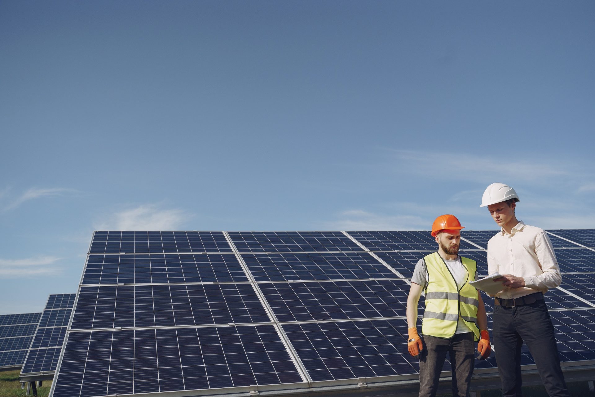 From below of serious young male colleagues in helmets working at solar energy station against cloudless blue sky